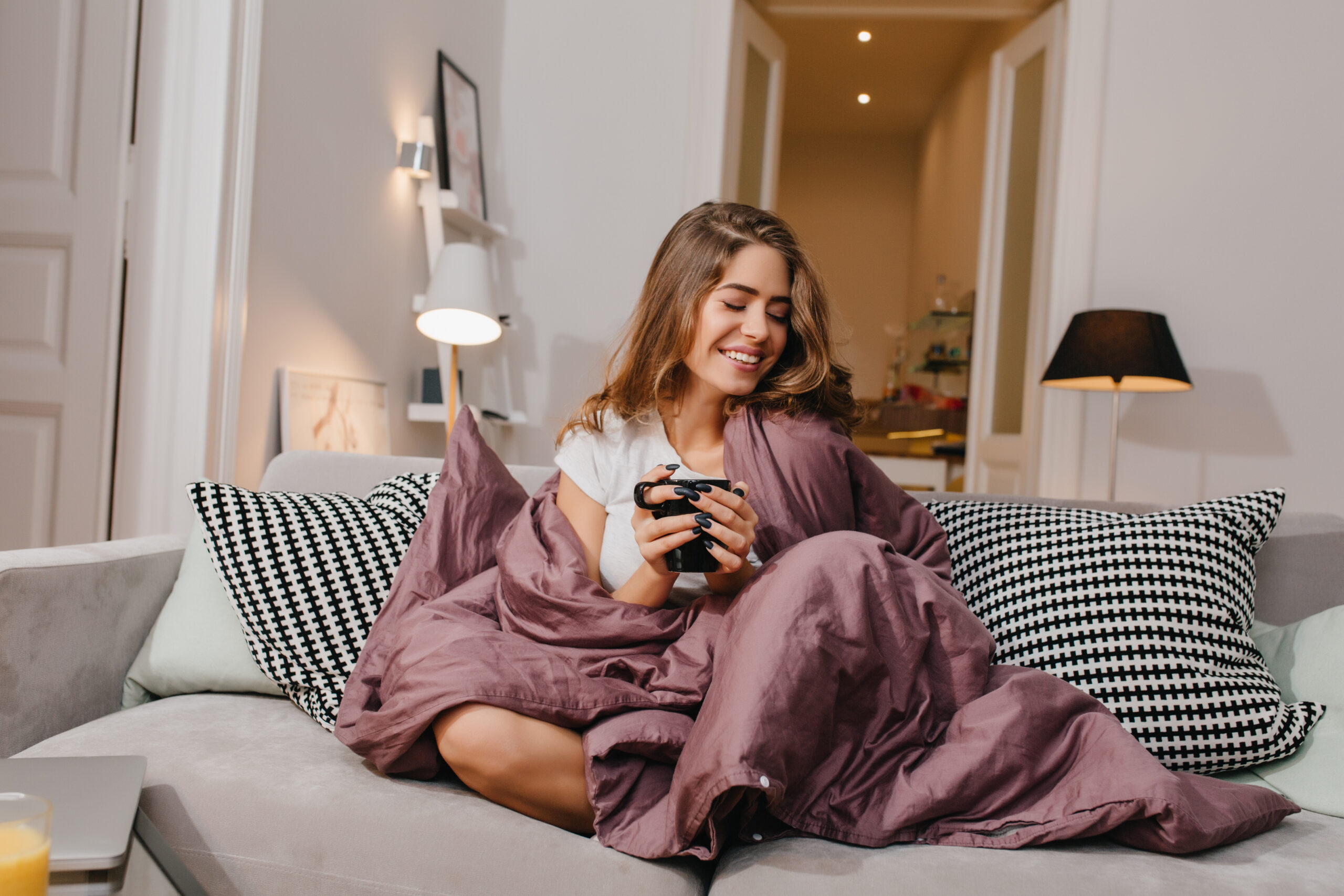 Cheerful girl sitting on couch with blanket and cushions and smiling. Spectacular brunette lady laughing, while drinking coffee under plaid.